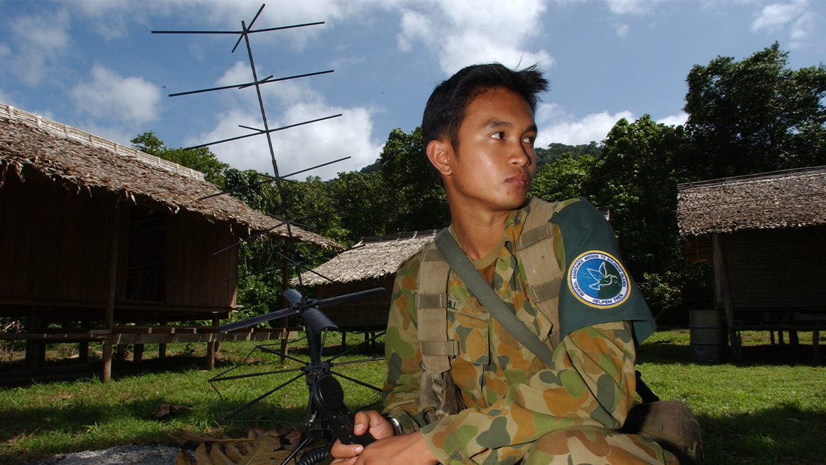 Soldier holding an antenna with straw roof huts in background