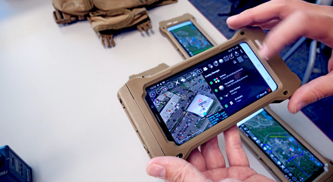 This colour photograph shows a small digital device held in a person’s hands. On the screen of the device can be seen satellite imagery, text and icons. On the table in the background are two more similar digital devices and a camouflaged carry pack.