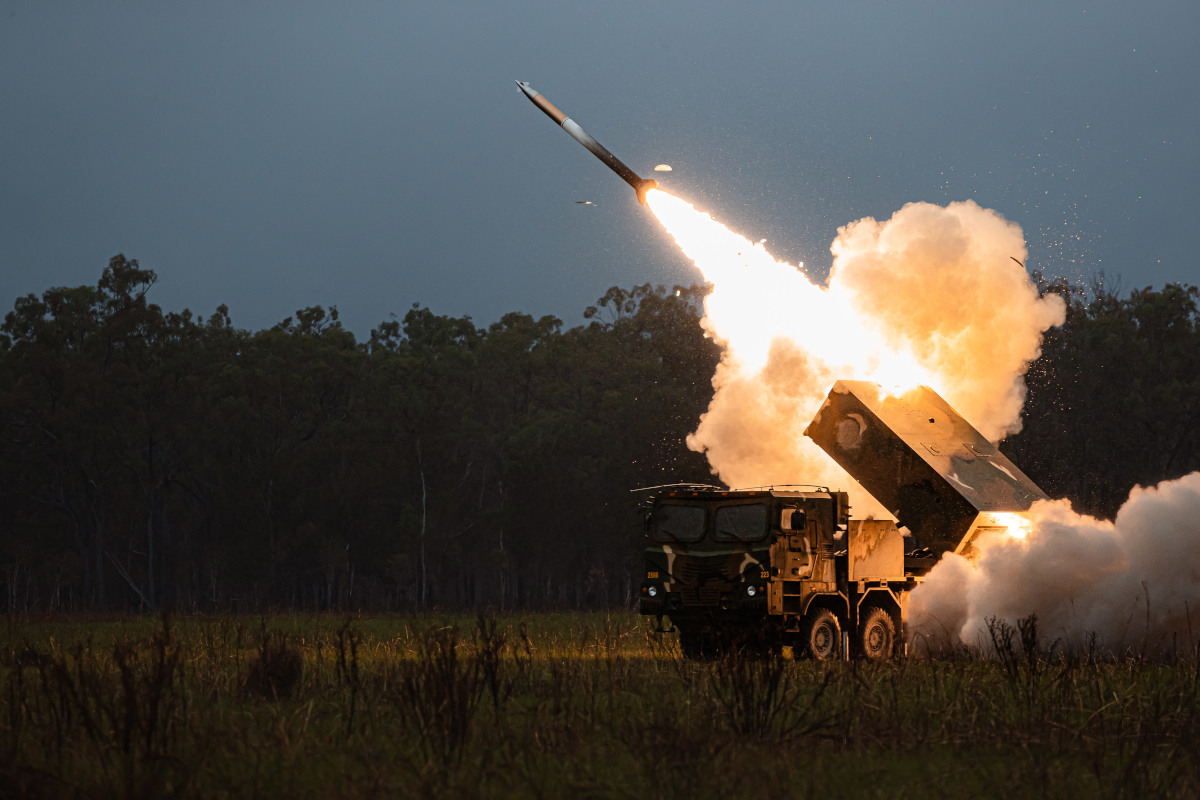 This coloured photograph shows a clearing in the Australian bush at night. Parked in the clearing is a military truck mounted with a multiple launch rocket system. A rocket can be seen launching from the system into the air, surrounded by smoke and light.