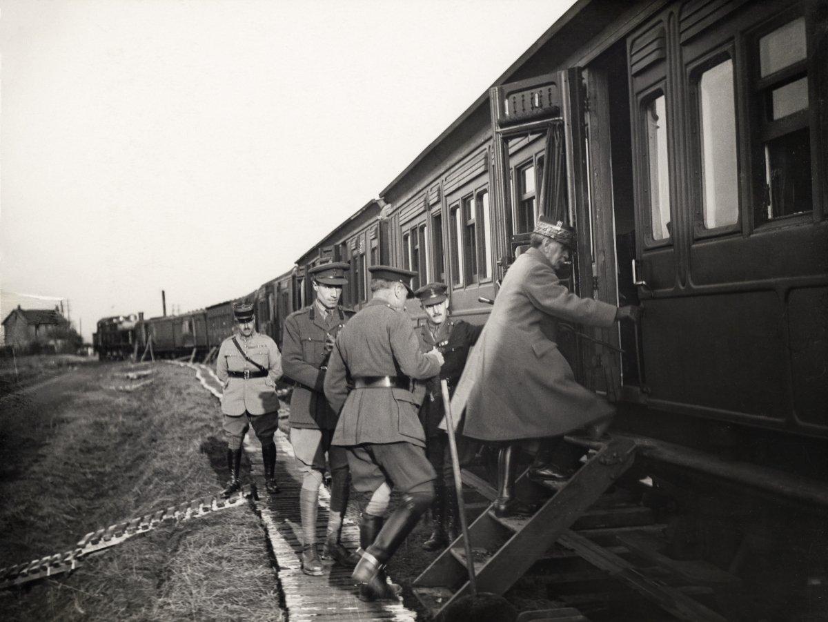 This black and white photograph shows a train stopped in a field next to an embankment. Wooden duckboards are seen on the ground parallel with the track, and leading up the embankment to the track. A small set of wooden steps lead up from the duckboards to the carriage, and several men dressed in Second World War military uniform and overcoats are seen boarding the carriage.