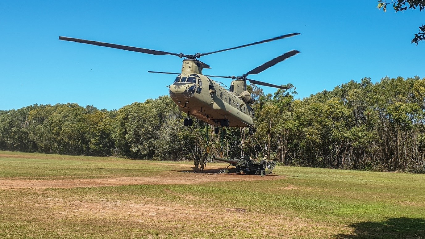 This colour photograph shows a grassed area with trees in the background in the middle of the day. A CH-47 helicopter hovers above the ground, and soldiers dressed in combat uniforms and armed with rifles can be seen unhooking an artillery gun from the tethers which attach the gun to the helicopter.