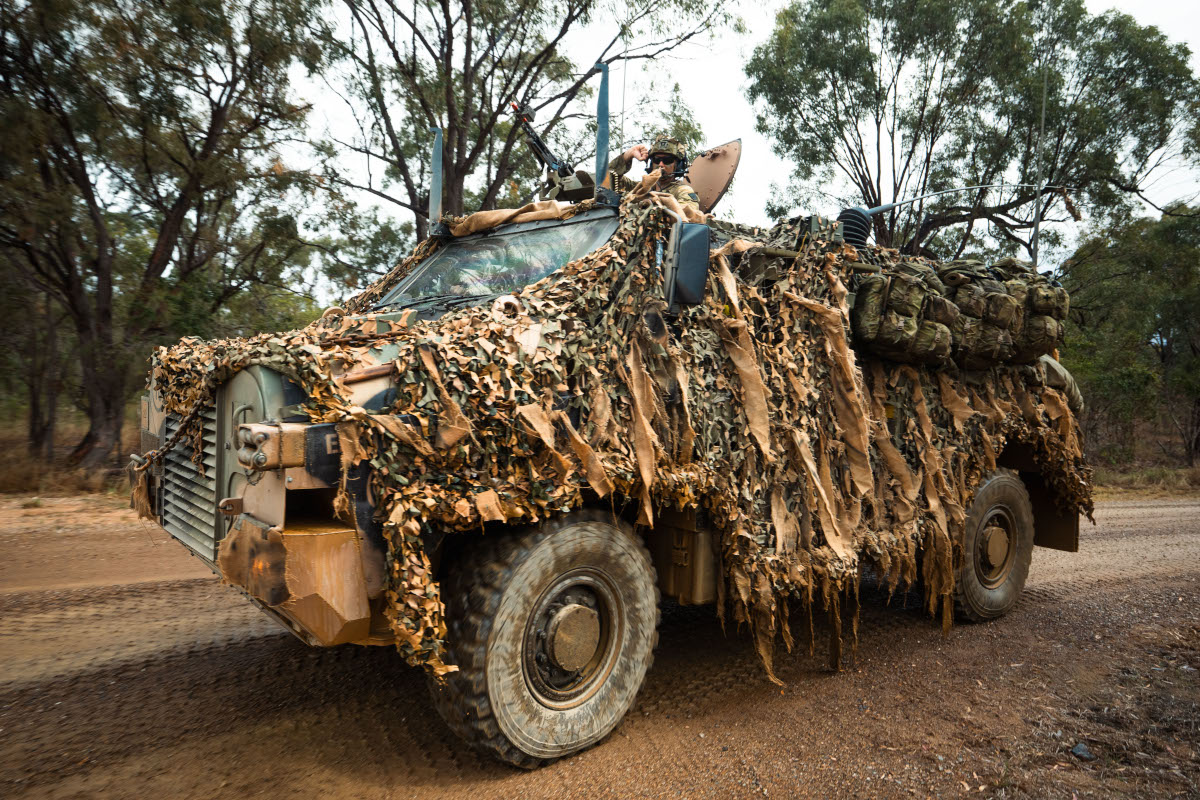 This coloured photograph depicts a military truck on a dirt road in the Australian bush. The truck is covered with camouflage material. Soldiers’ personal packs are mounted on the side. Several antennas can be seen protruding from the  top of the vehicle and a soldier wearing a helmet can be seen in a turret behind a machine gun.