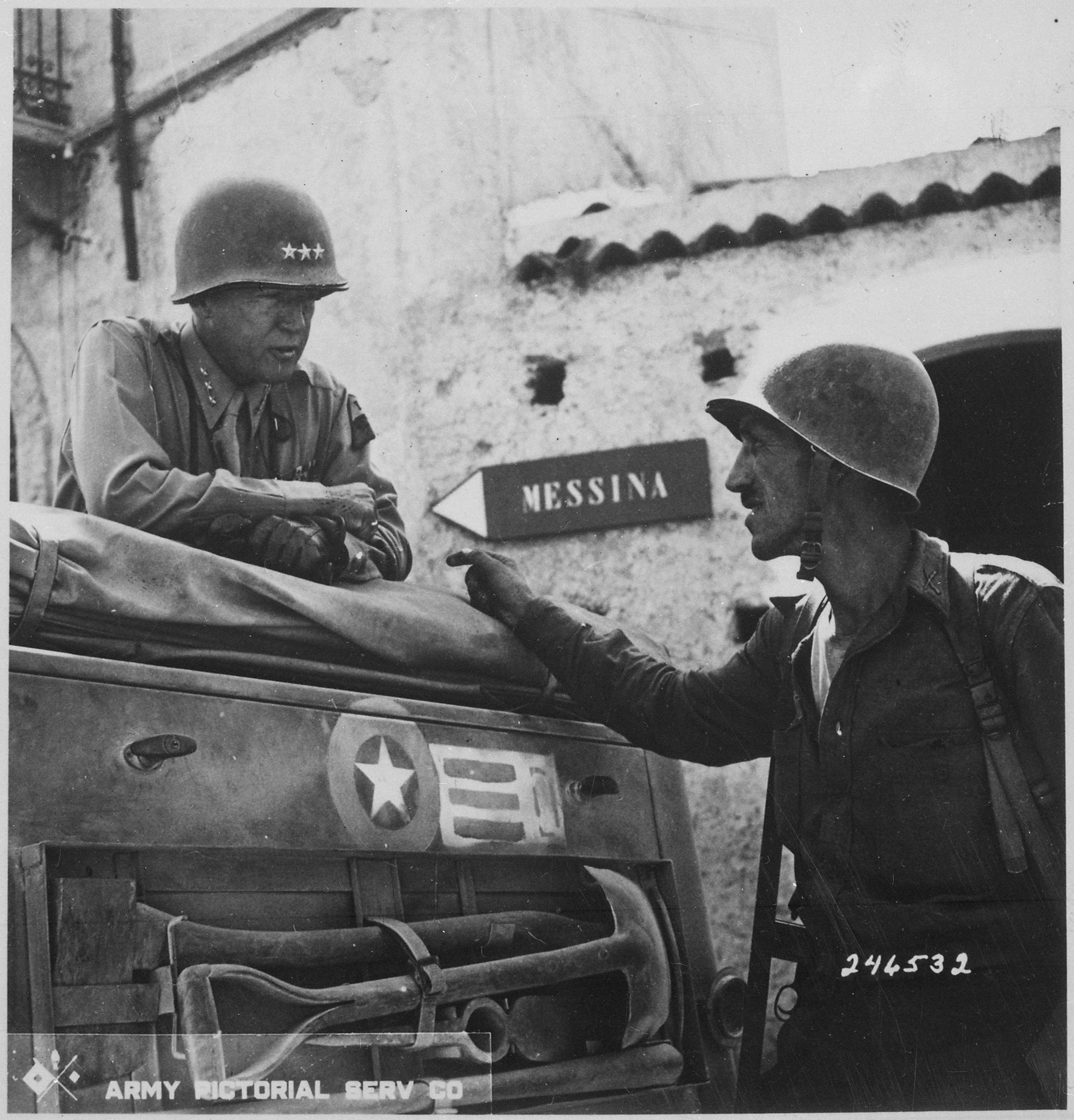 Lieutenant General George S. Patton confers with Lieutenant Colonel Lyle Bernard in a street near Brolo, Sicily on either 12 or 13 August 1943. A sign on a wall in the background shows the directions to Messina.