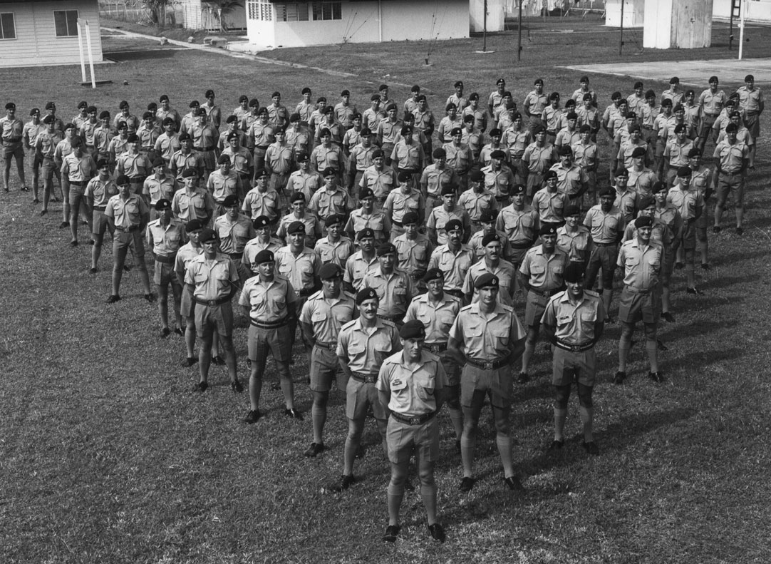 Soldiers from a Rifle Company Butterworth deployment in the 1980’s are shown gathered in a triangular formation for an official photograph. The soldiers are wearing Australian Army polyester barracks dress and berets. They are standing at ease in a grassy area with buildings in the background. The group includes General Angus Campbell (then a lieutenant).