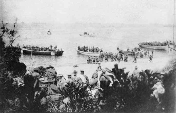 Several long row boats containing troops disembark onto a beach observed by other soldiers watching from within low vegetation