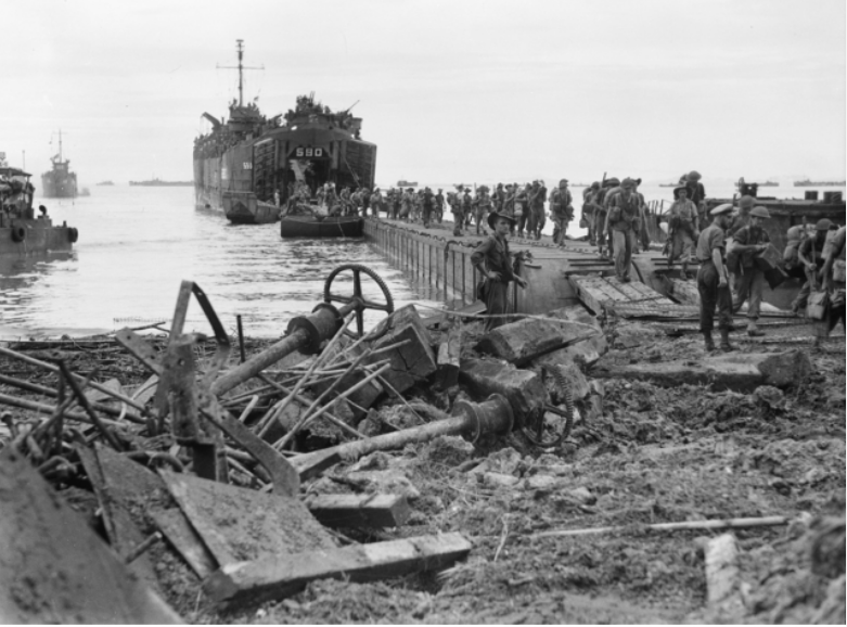 Australian troops disembarking Landing Ship Tank (LST) No 590 at Tarakan. LST 590 dominates the middle of the photograph and is tied up at one end of pontoon bridge that connects the ship to the shore. Australian troops are making their way from the vessel, over the pontoon bridge to the shoreline, on the right side of the image.