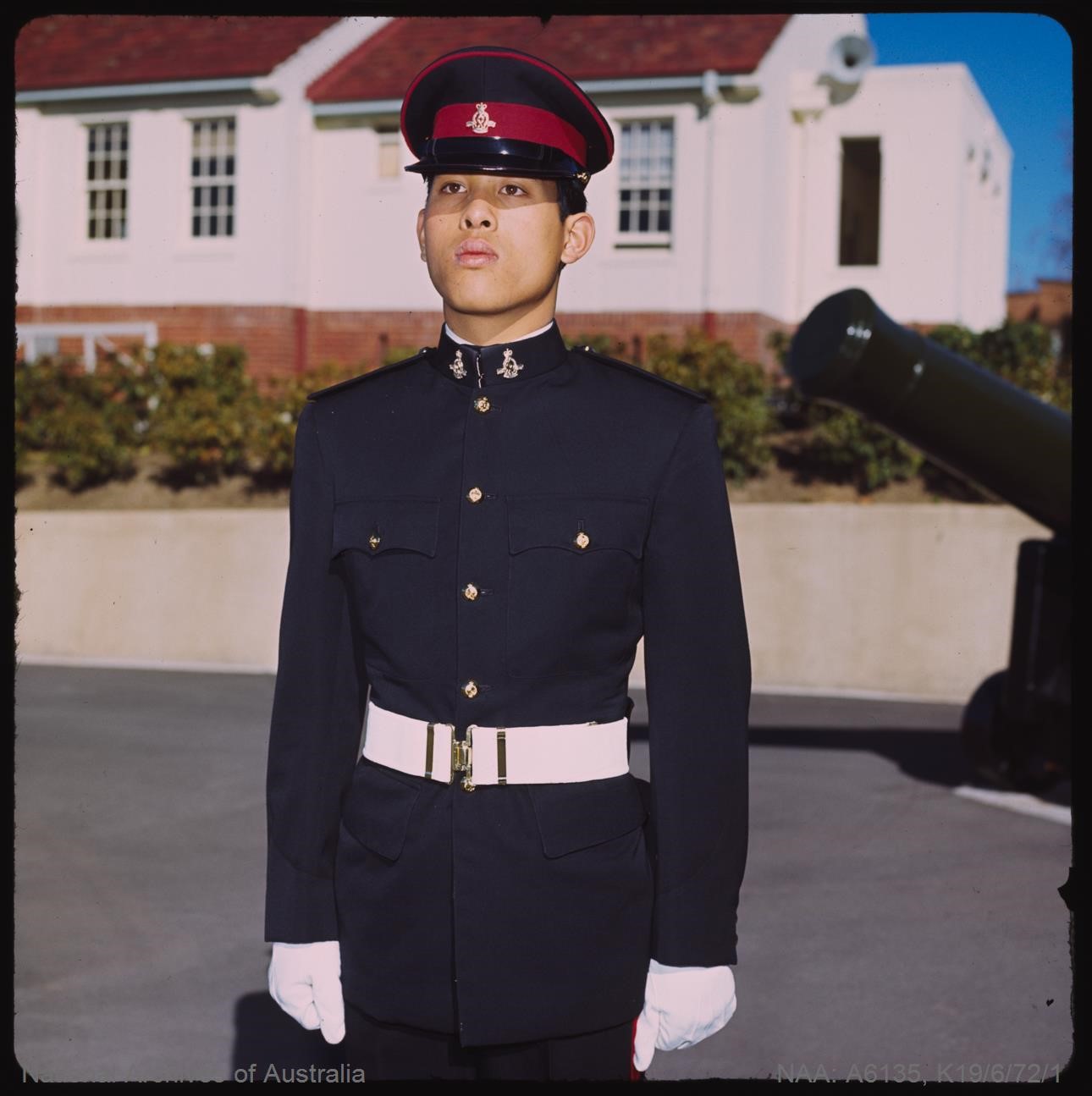 His Royal Highness the Crown Prince of Thailand – Vajiralongkorn Mahidol is shown while he was a cadet at the Royal Military College Duntroon in 1972. He is standing to attention on the parade ground, flanked by an old muzzle loading cannon, with white washed accommodation buildings behind him. He is wearing Patrol Blues ceremonial uniform including a blues peak cap, white parade belt and white gloves.