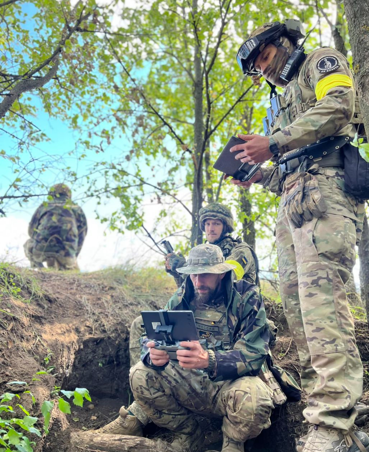 Soldiers in a lightly wooded area on a small dirt rise, two soldiers looking at tablets.