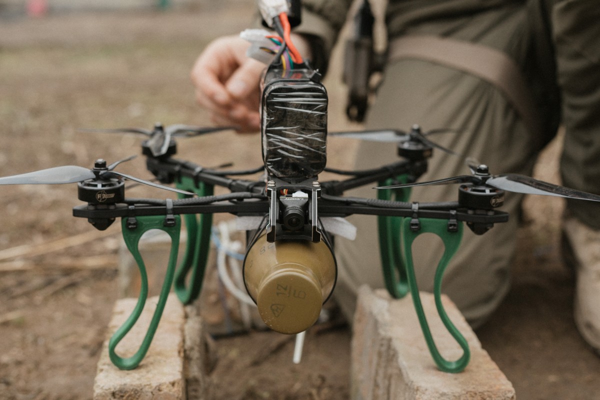 Multi-rotor drone with explosive attached with tape; soldier on one knee, crouching behind drone.