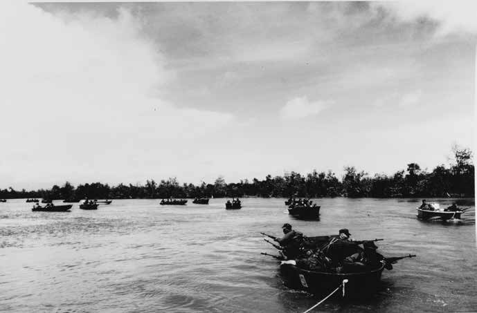 Several military landing craft with armed military personnel on board approaching a coastline fringed by tropical vegetation.