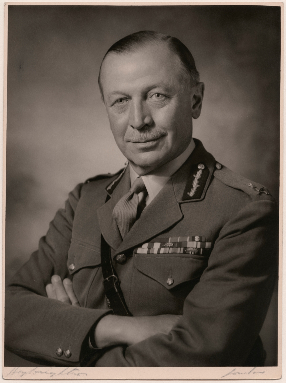 A black-and-white studio portrait showing a uniformed military officer seated with arms folded. The officer is wearing a decorated military tunic with visible insignia and medal ribbons. The photograph is presented within a narrow rectangular frame against a plain background. Caption text beneath the image identifies the subject as Sir John Hackett and notes that the photograph was taken by Hay Wrightson Ltd. in the 1960s.