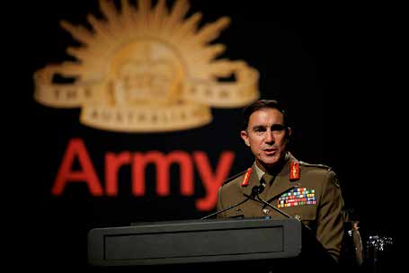 A uniformed Australian Army officer stands at a lectern delivering a presentation at a formal Army event. The officer is wearing a service uniform with rank insignia, ribbons and decorations. In the background, a large Australian Army emblem and the word “Army” are prominently displayed on a dark stage backdrop. The image is taken from the audience perspective and focuses on the speaker at the podium during an address at the Chief of Army Symposium 2024 in Melbourne, Victoria.