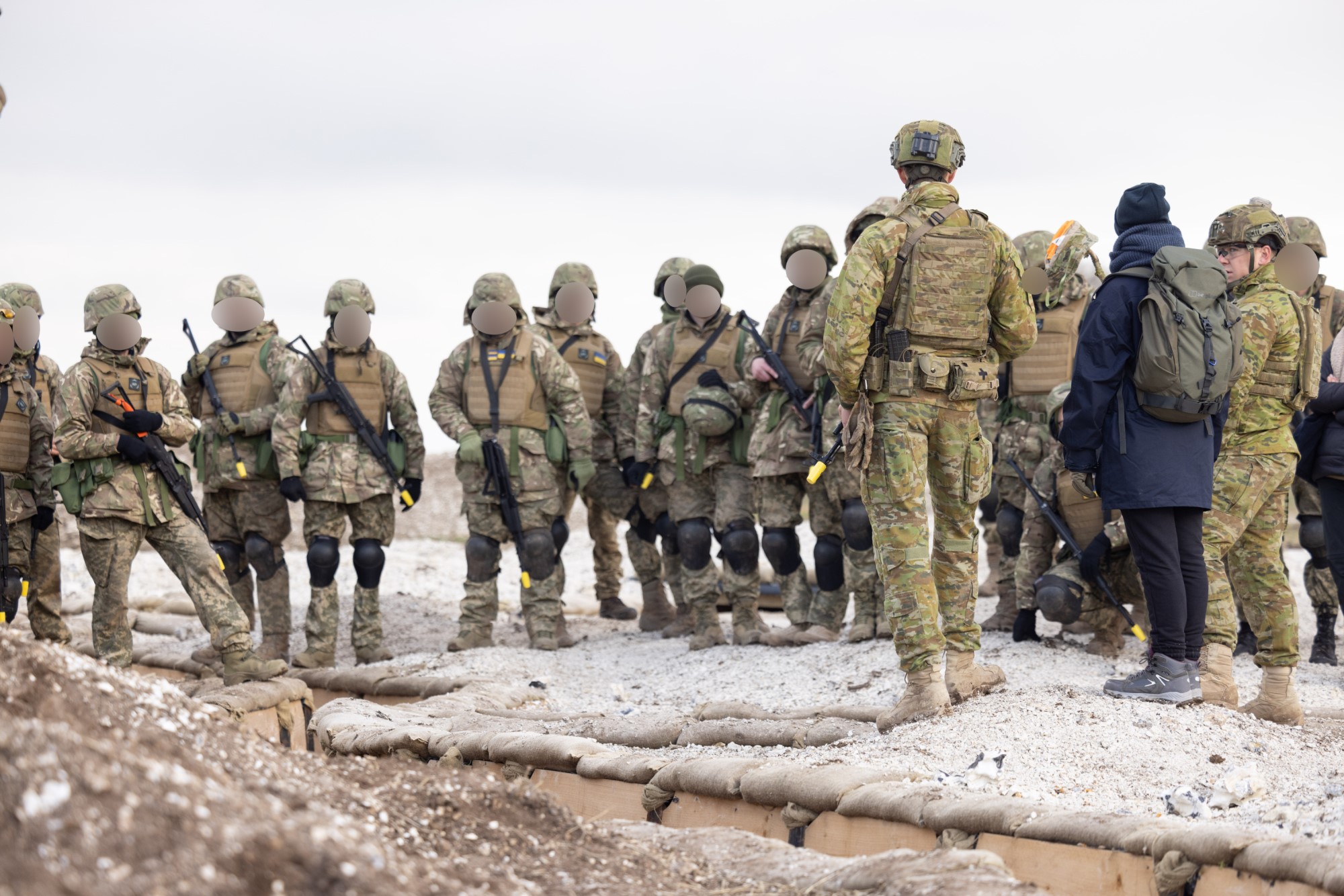 Approximately 15 Ukrainian Army trainees wearing combat clothing, helmets, body armour and are carrying weapons in their hands or slung across their chests; distributed left to right across the mid-ground of the image on the far side of a trench. Trench extends from right foreground, to left background. A civilian dressed person with two Australian soldiers in right foreground wearing Australian uniforms (including cold weather jackets), combat helmets, load carrying equipment and combat body armour.