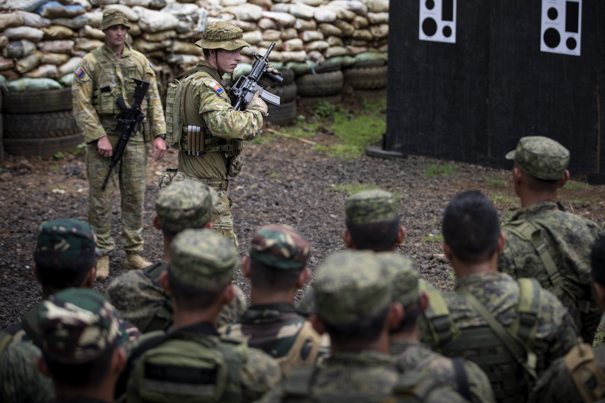 Two Australian soldiers in mid-ground of photograph wearing Australian uniforms, , one soldier is holding an AR 15 assault rifle half turned to the right. The other Australian soldier at the rear with weapon slung across chest. A group of Philippines Armed Forces in foreground facing the Australian, away from the camera. They are wearing military camouflage uniforms. They are standing on a patch of gravel and at the rear of the photograph is a sandbag wall.