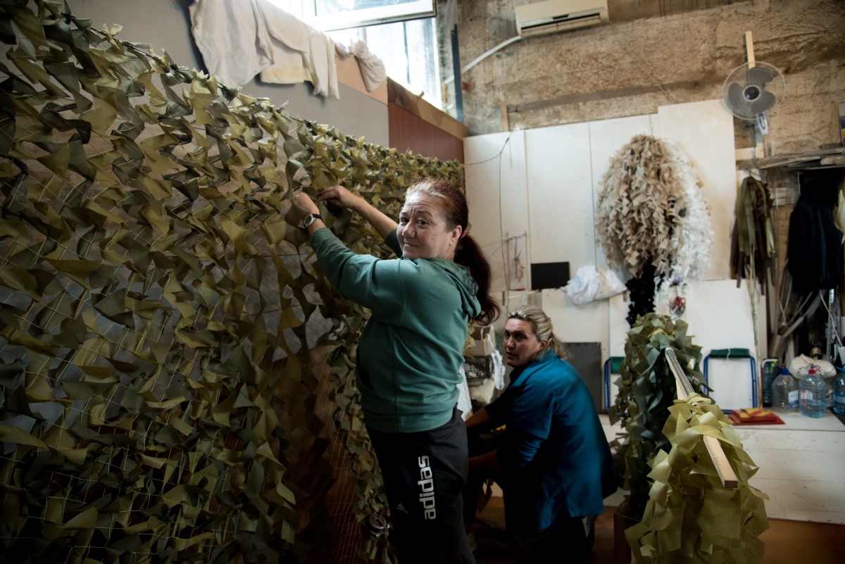 Two women work together in a workshop, attaching strips of fabric to a large camouflage net. The space around them is filled with materials, tools, and additional nets in progress.