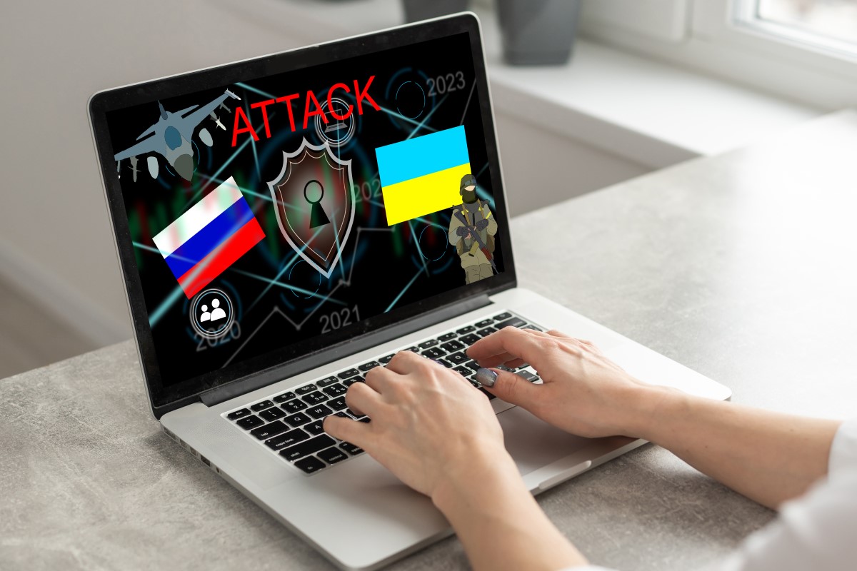 Two hands are shown typing on a laptop displaying graphics related to a cyberattack, including the Russian and Ukrainian flags, digital network icons, and the word “ATTACK” in red across the top of the screen.