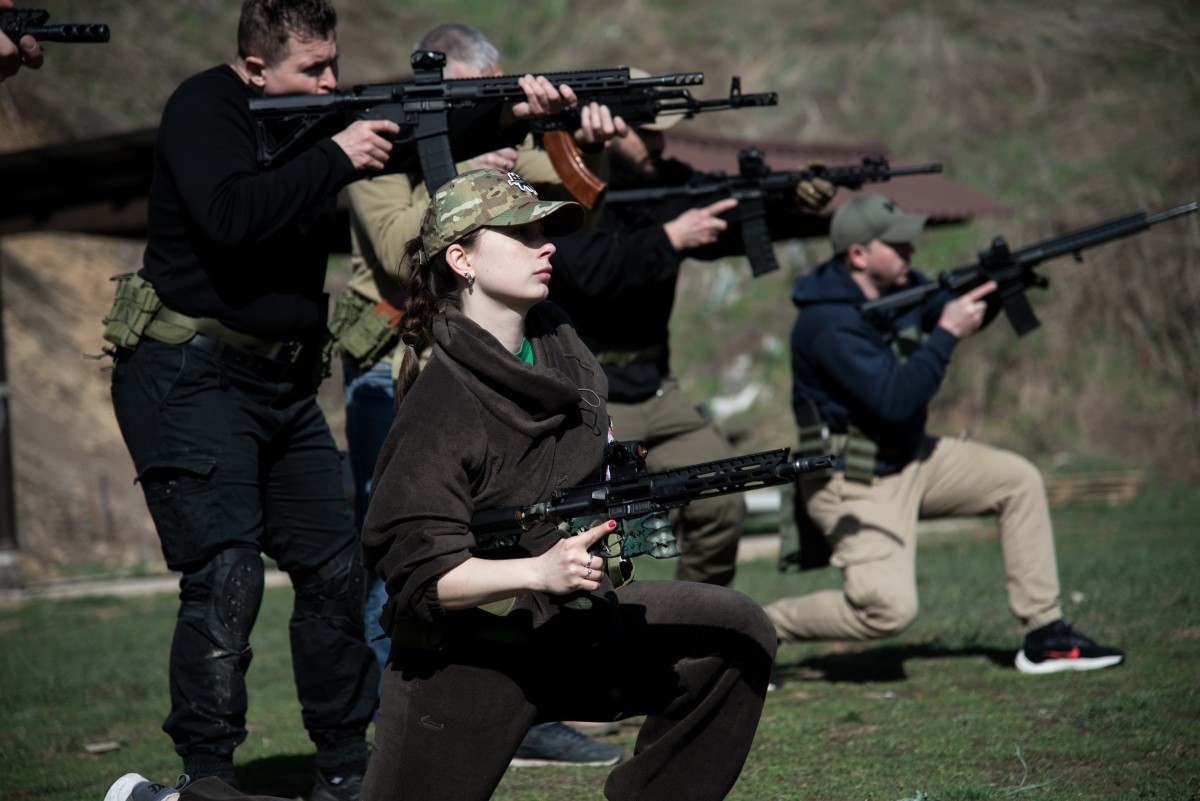 A group of people kneel and stand in a staggered formation outdoors. They are dressed in a range of civilian attire including camouflages and plain forage caps. They are armed with a variety of military assault rifles and are carrying out firing drills during a firearms training exercise.