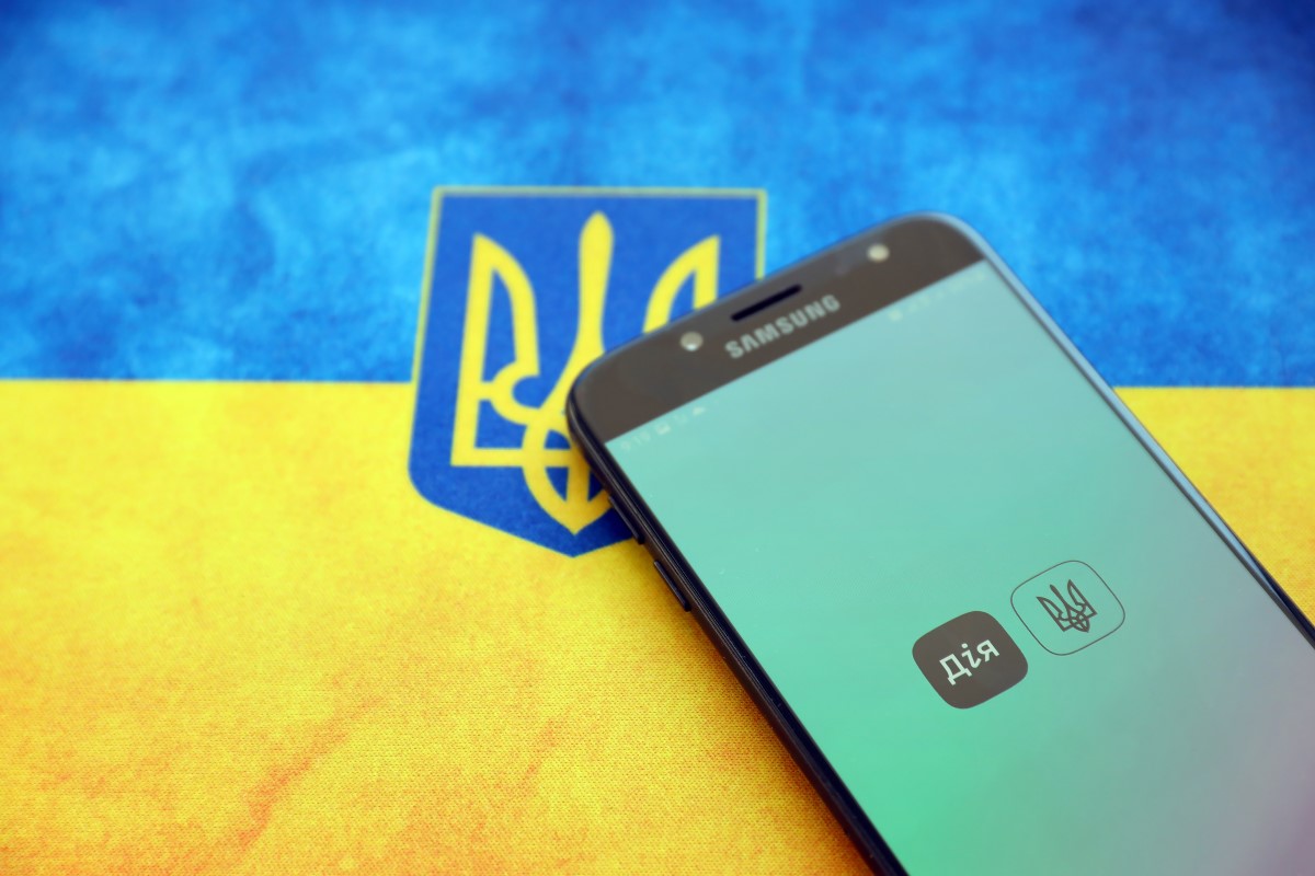 A smartphone displaying the Diia app screen rests on a surface in front of a Ukrainian flag featuring the national trident emblem.