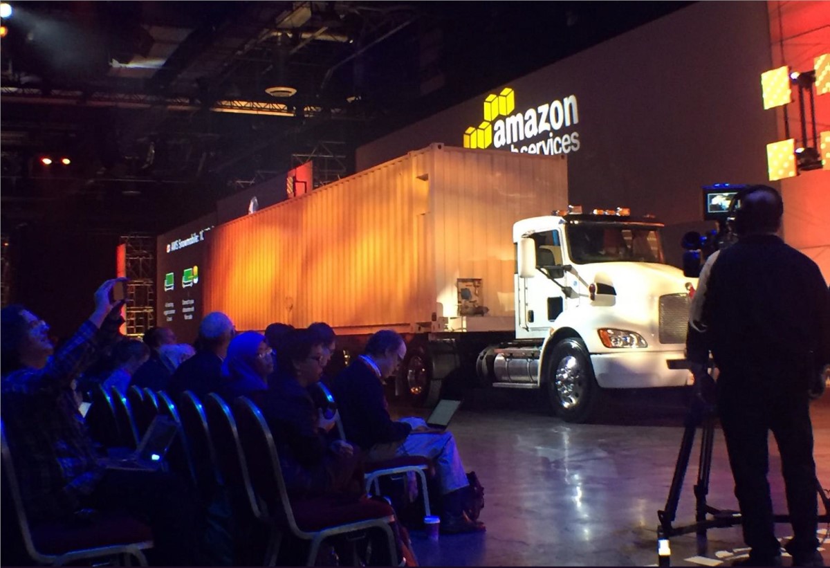 A large white truck and shipping container are displayed on stage at a transport industry event, with an audience seated in front and a camera operator filming. The backdrop features the Amazon Web Services logo.