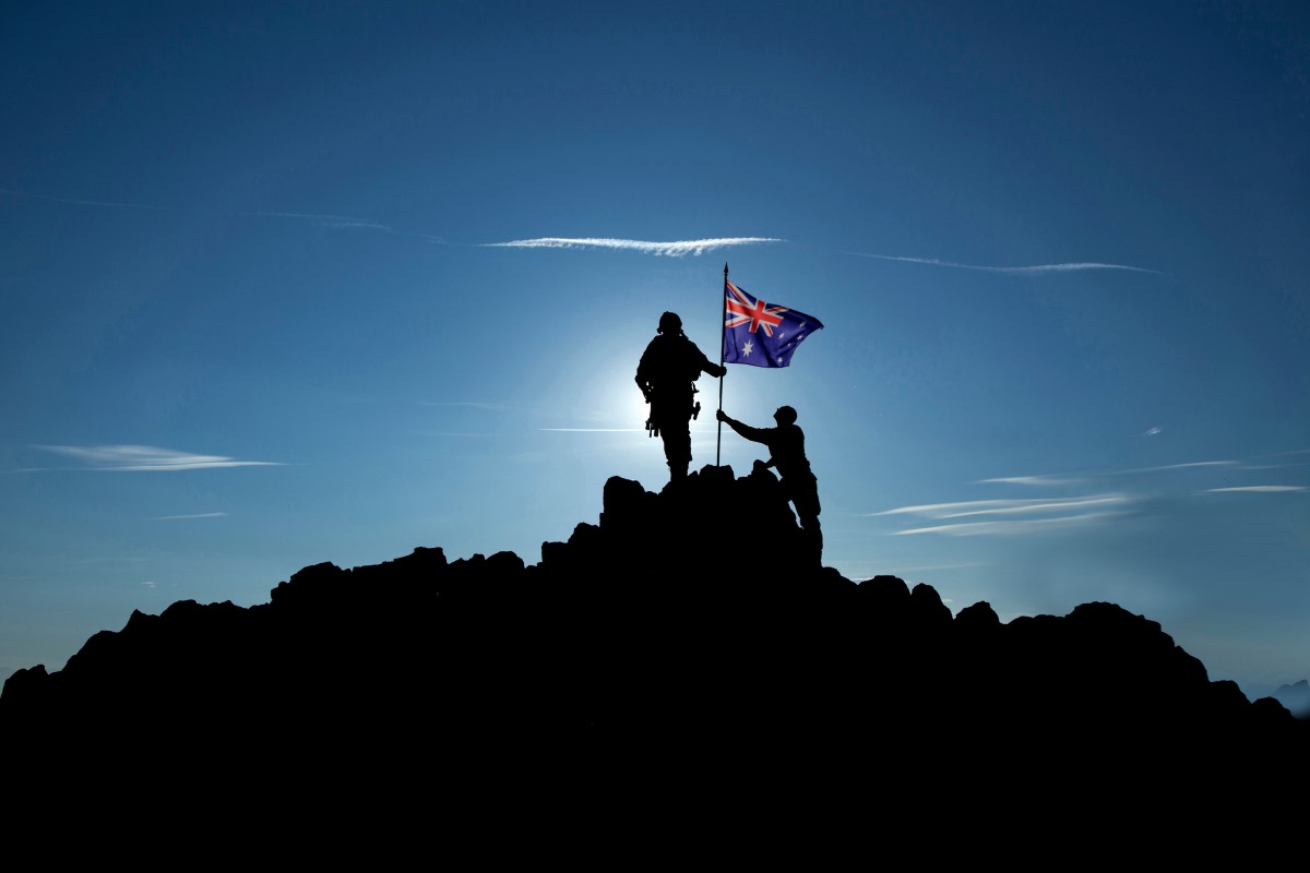 Two military figures are shown silhouetted against the rising sun on top of a rocky outcrop. The figures and the mountain top are in a deep shadow and no other features can be discerned. They are in the process of raising the Australian national flag on a fixed flag pole.