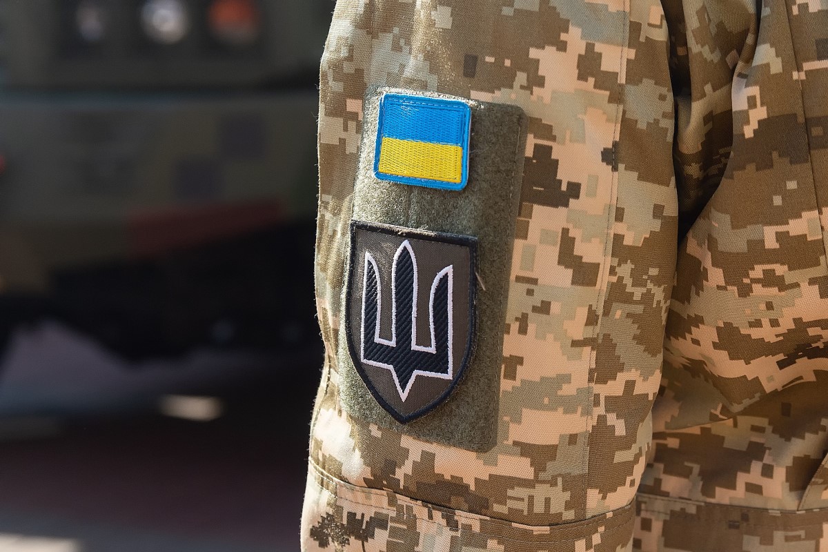 The image shows a close up of the left shoulder of a person dressed in Ukrainian camouflage uniform. The Ukrainian national trident symbol in black with white outlining on a shield is shown underneath a blue and yellow rectangle.