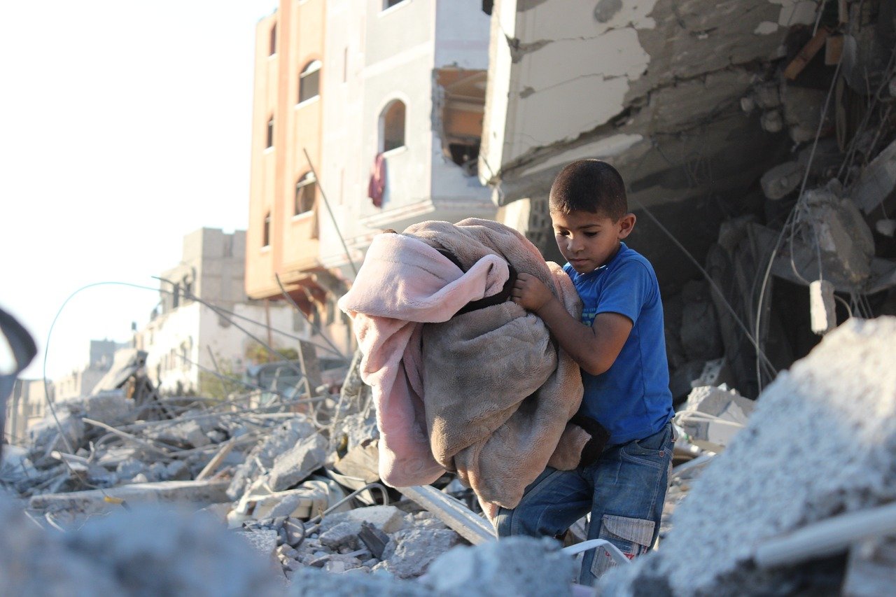 A child carrying blankets through rubble from destroyed building in Palestine Gaza Strip. Image by hosny salah from Pixabay.