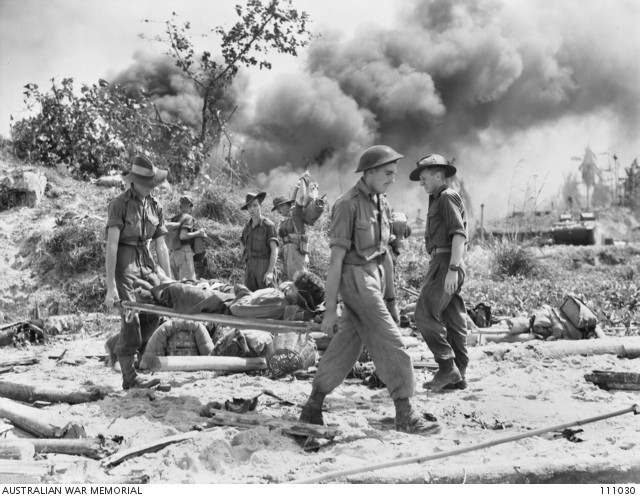 Stretcher-bearers carry a wounded man back to the dressing station set up on the landing beach at Balikpapan by troops of the 2nd Beach Group, 01 Jul 1945. Behind them, other unit members set up a temporary advanced headquarters to control beachhead operations.