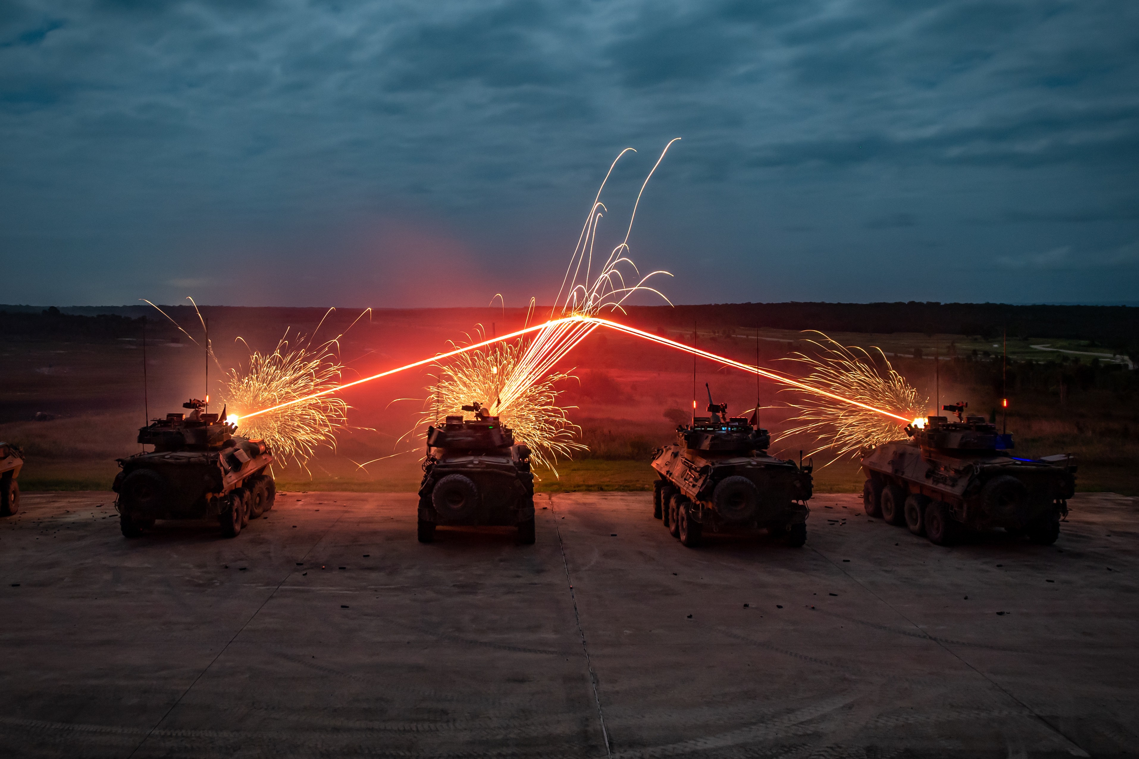 Four armoured vehicles in a row firing at a central point in semi-darkness; firing has produced red lighting effect