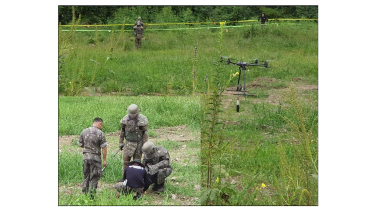 Soldiers in a green field with irregular long grass. Group of three are investigating the ground with one multi-rotor uncrewed aerial system is airborne to their right.