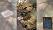 A soldier crouching and holding a 3D printed tile with a camouflage net in the background and with the cover of Spotlight Brief 7/21 in the foreground lower right corner