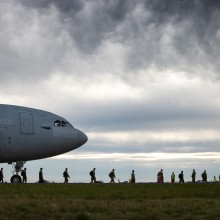 Nose of military transport aircraft from left with troops along ground walking away from the aircraft in single file