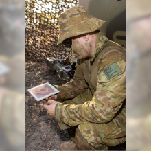 A soldier crouching and holding a 3D printed tile with a camouflage net in the background and with the cover of Spotlight Brief 7/21 in the foreground lower right corner